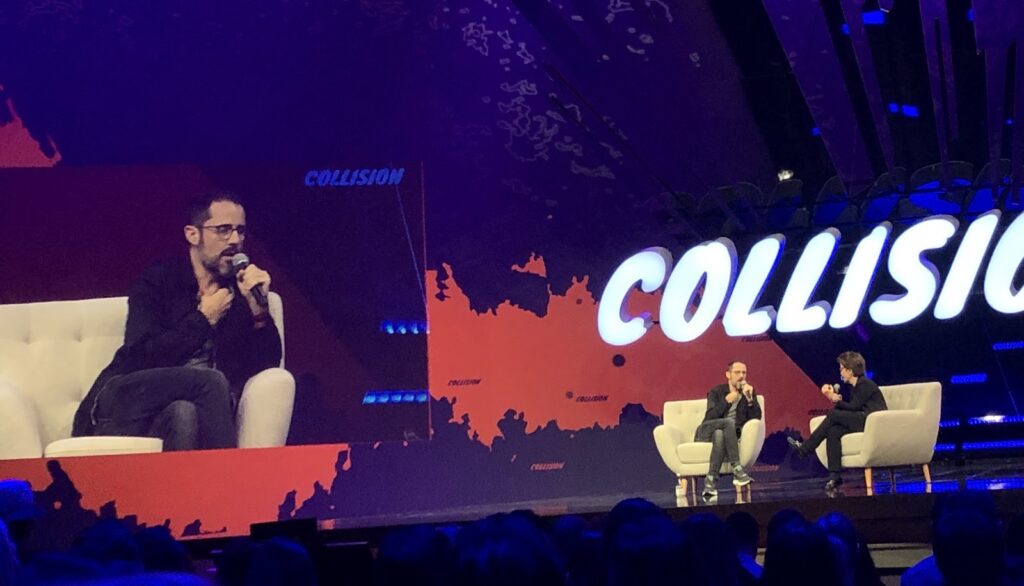Two speakers seated on stage in armchairs during a talk at the Collision conference, with a large "COLLISION" sign in the background.