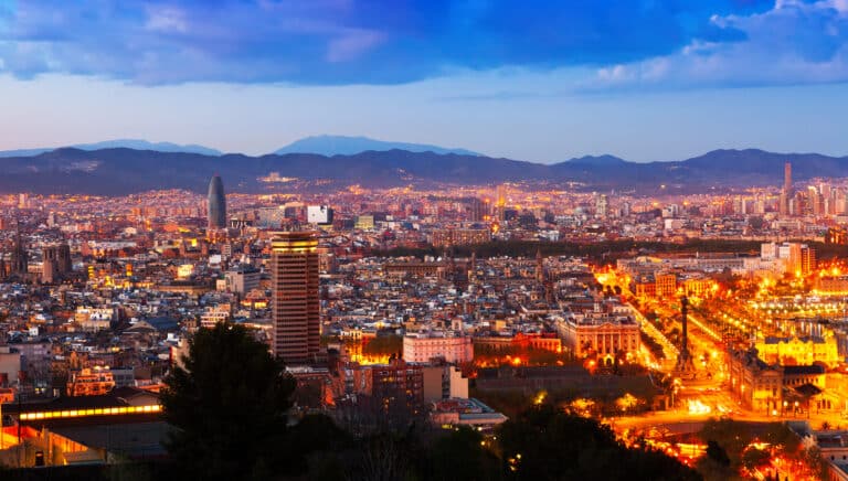 A panoramic view of Barcelona at dusk, showcasing illuminated buildings and vibrant city lights against a backdrop of mountains.