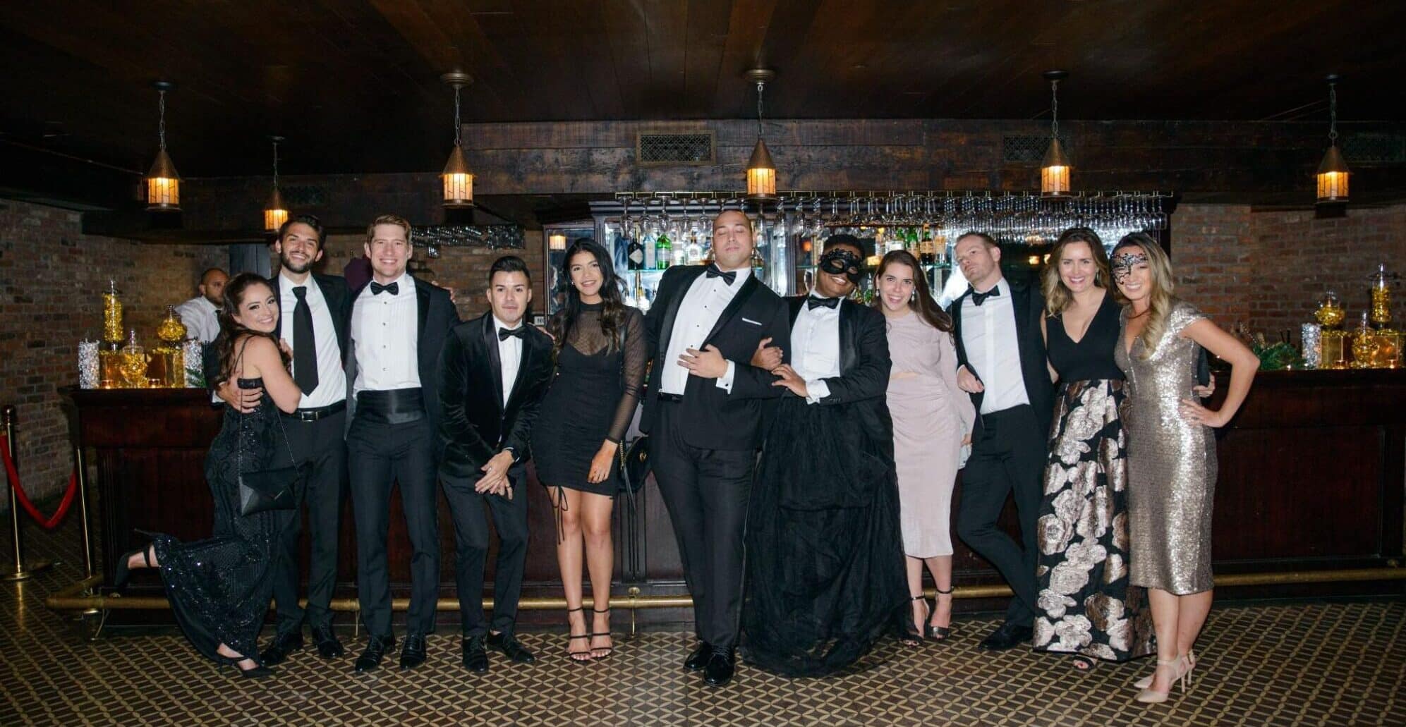 A group of people in formal wear pose at a stylish indoor event with a bar and warm lighting in the background.