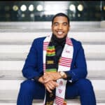 Man in a blue suit sitting on steps, smiling, wearing a multicolored Kente graduation stole