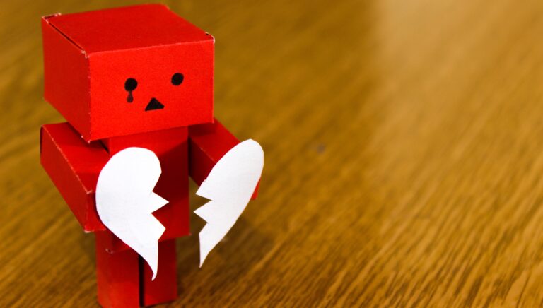 A red paper robot holding a broken white heart, expressing sadness, on a wooden surface.