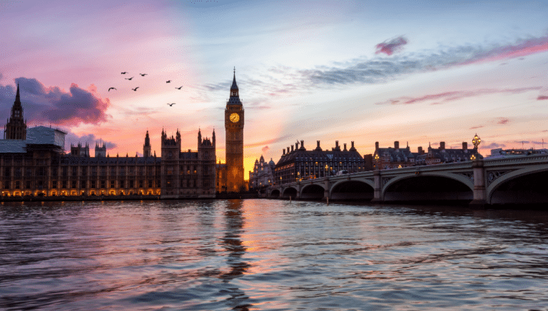 Sunset over Thames River with silhouette of Big Ben and Westminster Bridge, birds flying in the sky.