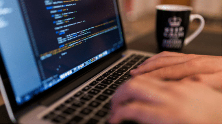Close-up of hands typing on a laptop with code on the screen and a coffee cup beside it.