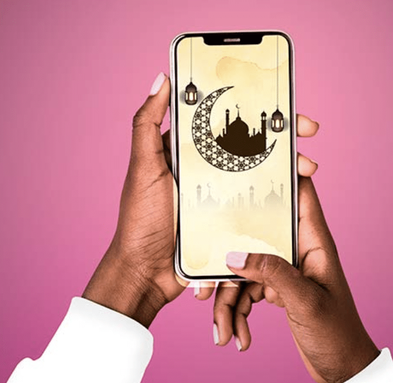 A hand holds a smartphone displaying a crescent moon and mosque silhouette against a soft pink background, symbolizing spirituality.