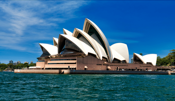 Sydney Opera House with white tiled shells, blue sky, and water in the foreground.
