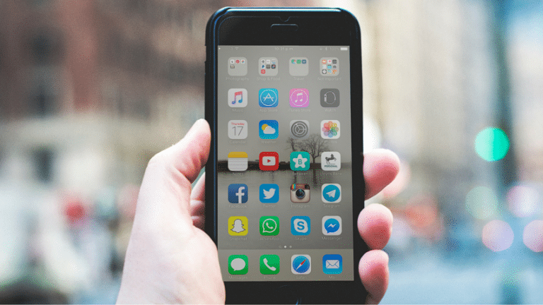 A hand holding a smartphone with various app icons on the screen, against a blurred city background.