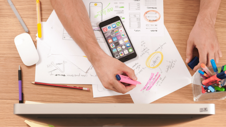 Person highlighting notes beside a smartphone with various charts and a computer on desk.